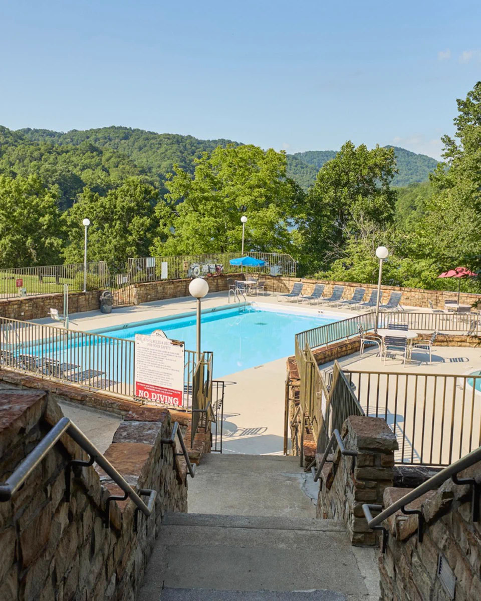 Outdoor swimming pool surrounded by a metal fence and situated among lush green trees and rolling hills. There are lounge chairs and umbrellas placed around the pool area. A "No Diving" sign is visible on one side of the pool.