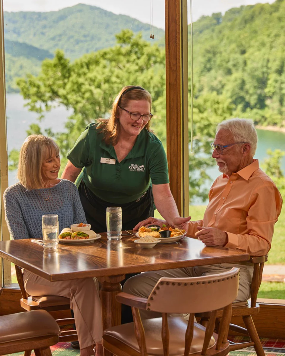 A group of three people are in a restaurant with large windows showcasing a scenic view of mountains and a lake. The server is handing a plate to one of the seated patrons. The diners have plates of food and glasses of water on the table.