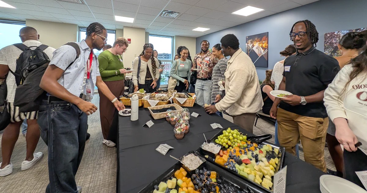 A large group of people is gathered around a buffet table with various foods, including fruit trays and snacks, in a modern room with artwork on the walls. Individuals are talking, smiling and serving themselves.