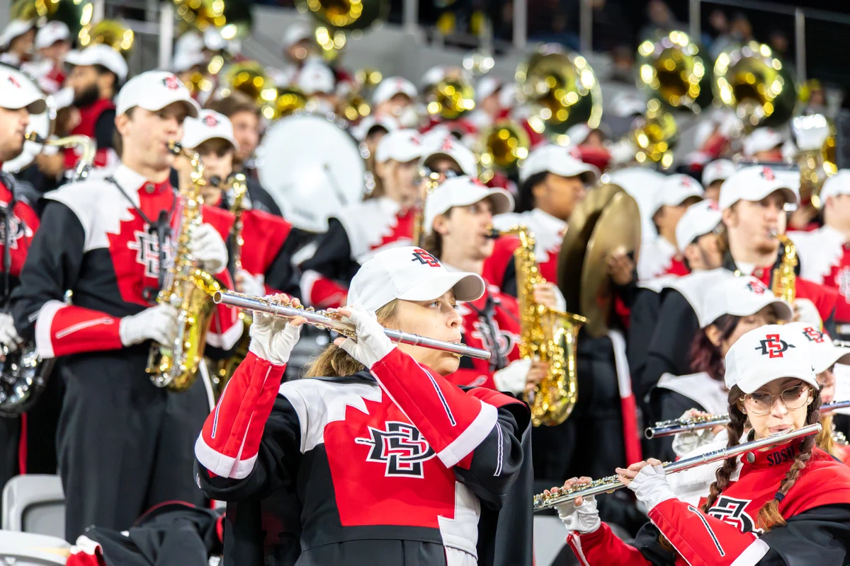 A marching band is performing at an outdoor event. Band members are playing various instruments, including flutes, saxophones, and tubas. They are wearing uniforms with "SDSU" logos and red, black, and white colors, along with matching caps.