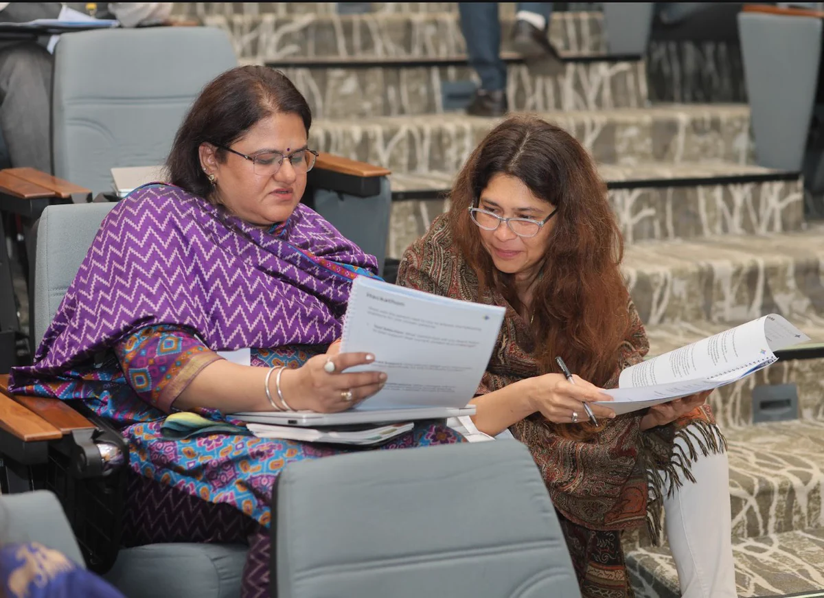 Two women seated in an auditorium, intently reviewing and discussing printed workshop documents together.