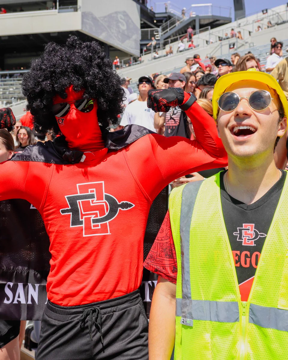 A person in a vibrant San Diego State University costume poses energetically with arms flexed. Next to them, another person wears a yellow safety vest, a yellow hard hat, and round sunglasses. In the background, a crowd of people enjoy a lively outdoor event.
