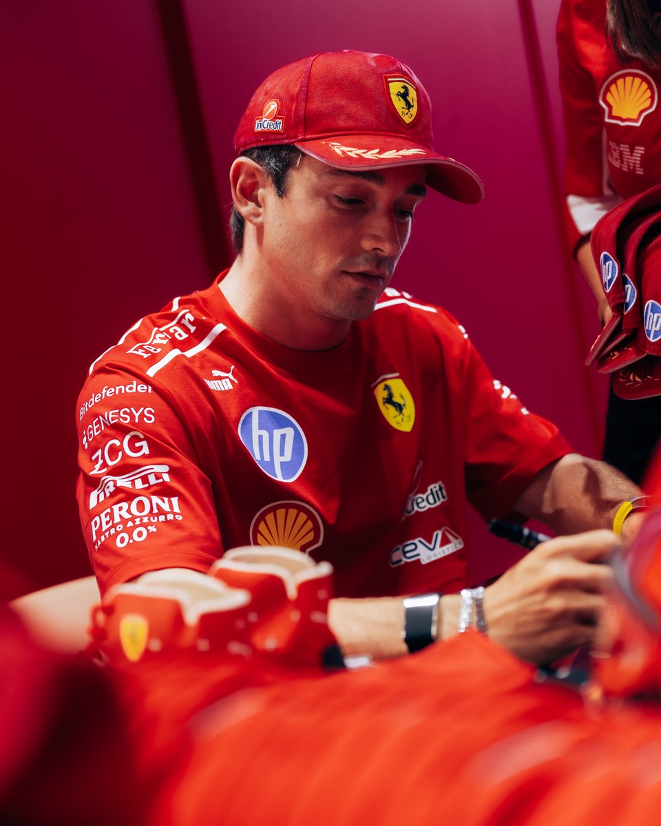 F1 driver Charles Leclerc, in a red Ferrari cap (UniCredit, Ferrari logo) and team shirt (HP, Shell, Peroni, Puma, Bitdefender, Ceva), looks down, signing. His red shirt features many sponsor logos against a vibrant red background.