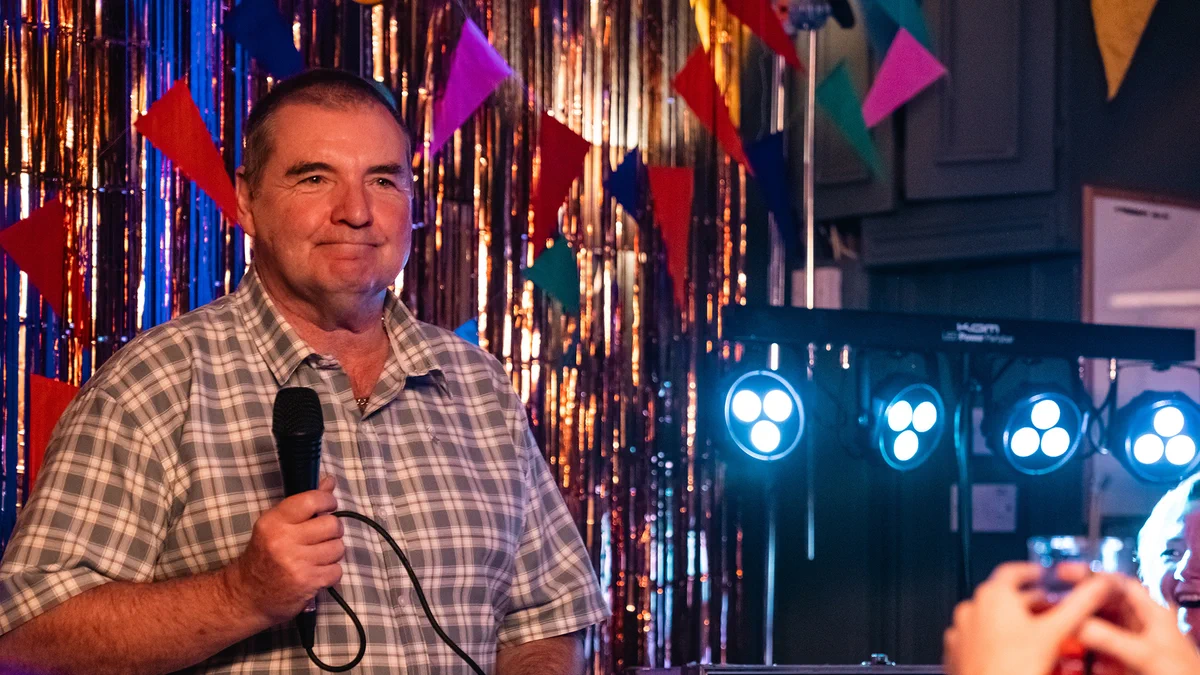 A man in chequered shirt stood holding a microphone smiling to a crowd at a party