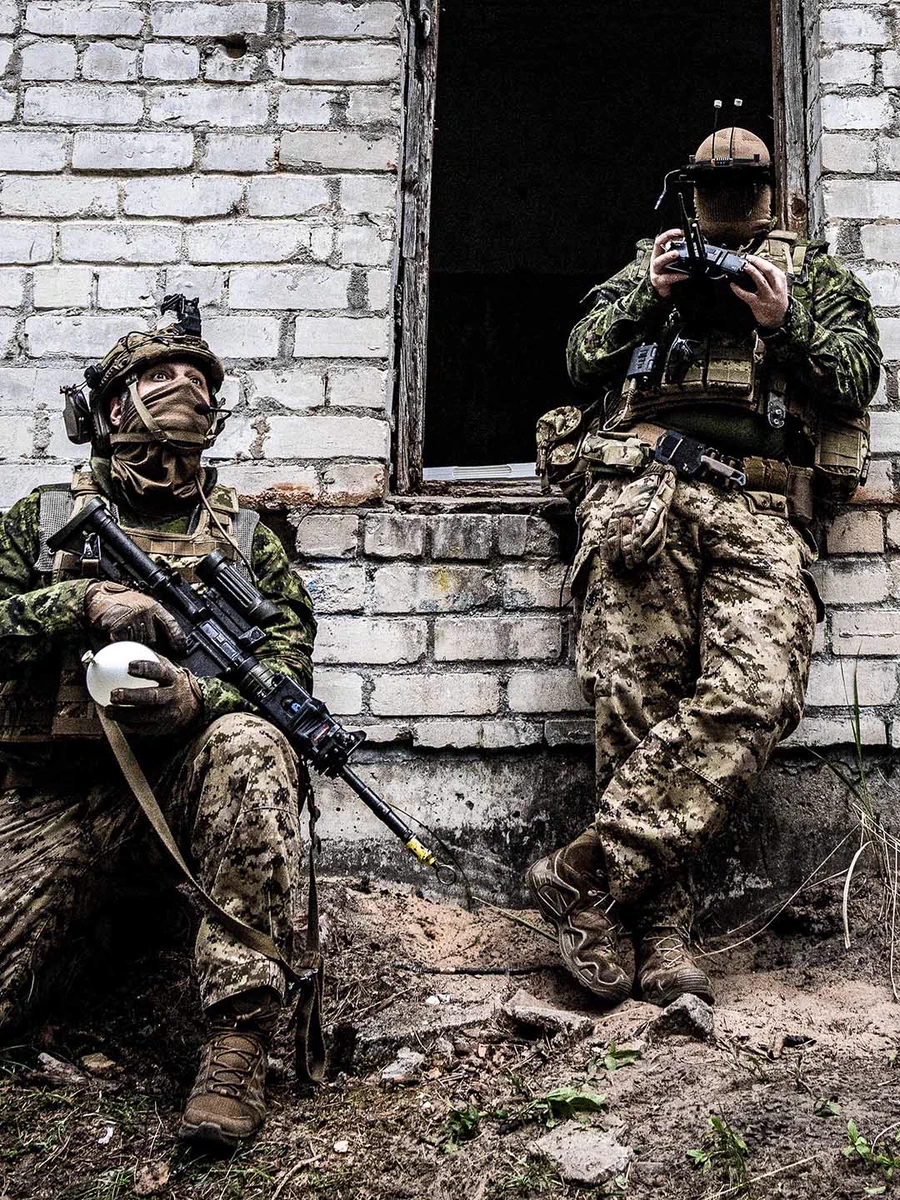 Two soldiers in camouflage gear during training, one holding a drone controller.