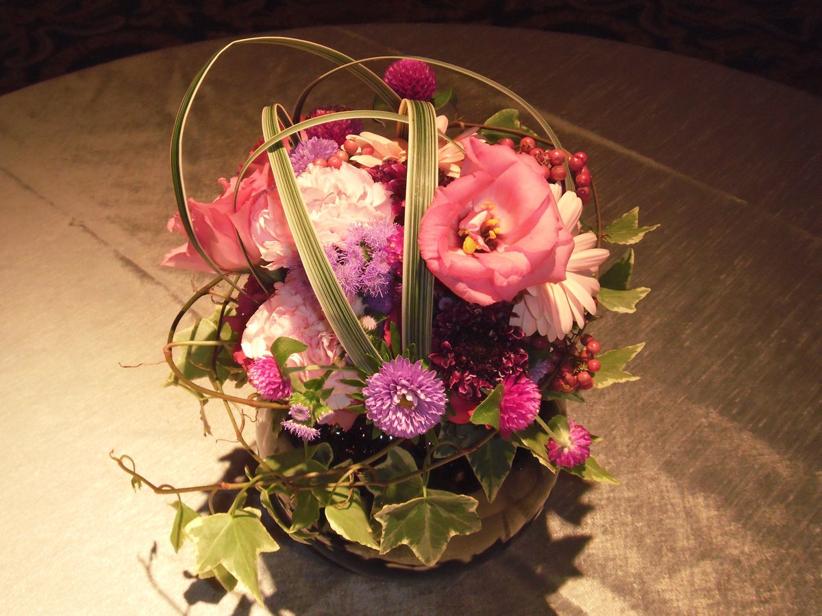 SS_tv_flower's tweet photo. 【生花と造花の使い分けは「画」と「温度」で決めています】

テレビの装飾を見て「これ生花ですか？造花ですか？」と聞かれることがよくあります。

バラエティ番組に限れば、体感で9割5分は造花です。ただ、常に造花というわけではなく、現場ごとに条件を見て決めています。

■判断基準1：カメラとの距離ではなく「画」

「カメラから遠ければ造花でいい」と思われがちですが、そうではありません。テレビカメラはそもそも距離を取った位置から撮り、ズームで画を作ります。カメラが5m離れていても、ズームで手元を抜くなら、その花は生花でないと持ちません。

見ているのは物理的な距離ではなく、その花が画のどこにどう映るかです。

・寄りで抜かれる卓上、出演者の手元 → 生花
・壁面装飾、高所、引きの画の背景 → 造花で成立
添付した写真の左が生花の卓上アレンジ、右が造花の壁面装飾です。壁面の方はシダの葉の質感まで作り込まれていて、少し離れれば見分けはつきません。

■判断基準2：撮影時間ではなく「温度と拘束時間」

こちらの方が判断を左右します。見るのは収録時間ではなく、美術トラックへの積み込みから搬入までを含めた総拘束時間です。

夏場の外ロケは基本的に造花です。撮影自体が1〜2時間でも、です。理由は2つあります。

ひとつは搬入までの時間で生花が弱ること。もうひとつが厄介で、水落ちが進むと、同じシーンの別カットを時間を空けて撮ったときに、ドラマの中では同じ時刻のはずなのに花だけしおれている、という事態が起こります。

逆に搬入のないスタジオセット（エアコン完備）なら、季節を問わず、水切れさせない限り照明下で一日持ちます。

目安になるのは美術トラック荷台の温度です。花の適温である15〜20度を保てる季節なら、搬入を伴うロケでも生花を使います。適温を保てない時期に現場で生花が必要な場合は、花材と花器を運んで現場で活けることもあります。

■実際の比率（すべて体感値です）

・バラエティ番組：生花0.5 : 造花9.5
・ドラマ全体：生花4 : 造花6
・ドラマのうちスタジオセット内：生花6 : 造花4

注目していただきたいのはドラマの2行です。同じドラマでも、撮影場所が変わるだけで比率が逆転します。この差分がそのまま、搬入と温度という条件の厳しさを表しています。
バラエティで造花が圧倒的なのは、壁面や高所の装飾が中心であること、番組のテイストに合わせた色指定があること、収録が長時間に及ぶことが理由だと考えています。
以前あった、生花の壺花を中央に置いて出演者が装花を挟んで対談する、というコンセプトの番組は今はほとんど見かけません。

生花と造花の使い分けは、センスや好みではなく、条件の読み合いです。

そして正直なところ、気温の高い日が長くなった昨今、生花を扱う立場としては年々厳しい環境になってきていると感じています。