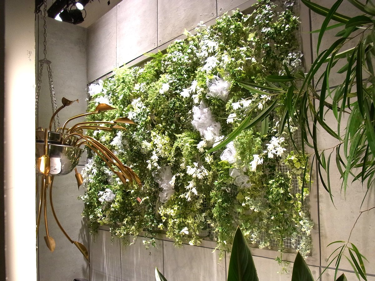 SS_tv_flower's tweet photo. 【生花と造花の使い分けは「画」と「温度」で決めています】

テレビの装飾を見て「これ生花ですか？造花ですか？」と聞かれることがよくあります。

バラエティ番組に限れば、体感で9割5分は造花です。ただ、常に造花というわけではなく、現場ごとに条件を見て決めています。

■判断基準1：カメラとの距離ではなく「画」

「カメラから遠ければ造花でいい」と思われがちですが、そうではありません。テレビカメラはそもそも距離を取った位置から撮り、ズームで画を作ります。カメラが5m離れていても、ズームで手元を抜くなら、その花は生花でないと持ちません。

見ているのは物理的な距離ではなく、その花が画のどこにどう映るかです。

・寄りで抜かれる卓上、出演者の手元 → 生花
・壁面装飾、高所、引きの画の背景 → 造花で成立
添付した写真の左が生花の卓上アレンジ、右が造花の壁面装飾です。壁面の方はシダの葉の質感まで作り込まれていて、少し離れれば見分けはつきません。

■判断基準2：撮影時間ではなく「温度と拘束時間」

こちらの方が判断を左右します。見るのは収録時間ではなく、美術トラックへの積み込みから搬入までを含めた総拘束時間です。

夏場の外ロケは基本的に造花です。撮影自体が1〜2時間でも、です。理由は2つあります。

ひとつは搬入までの時間で生花が弱ること。もうひとつが厄介で、水落ちが進むと、同じシーンの別カットを時間を空けて撮ったときに、ドラマの中では同じ時刻のはずなのに花だけしおれている、という事態が起こります。

逆に搬入のないスタジオセット（エアコン完備）なら、季節を問わず、水切れさせない限り照明下で一日持ちます。

目安になるのは美術トラック荷台の温度です。花の適温である15〜20度を保てる季節なら、搬入を伴うロケでも生花を使います。適温を保てない時期に現場で生花が必要な場合は、花材と花器を運んで現場で活けることもあります。

■実際の比率（すべて体感値です）

・バラエティ番組：生花0.5 : 造花9.5
・ドラマ全体：生花4 : 造花6
・ドラマのうちスタジオセット内：生花6 : 造花4

注目していただきたいのはドラマの2行です。同じドラマでも、撮影場所が変わるだけで比率が逆転します。この差分がそのまま、搬入と温度という条件の厳しさを表しています。
バラエティで造花が圧倒的なのは、壁面や高所の装飾が中心であること、番組のテイストに合わせた色指定があること、収録が長時間に及ぶことが理由だと考えています。
以前あった、生花の壺花を中央に置いて出演者が装花を挟んで対談する、というコンセプトの番組は今はほとんど見かけません。

生花と造花の使い分けは、センスや好みではなく、条件の読み合いです。

そして正直なところ、気温の高い日が長くなった昨今、生花を扱う立場としては年々厳しい環境になってきていると感じています。