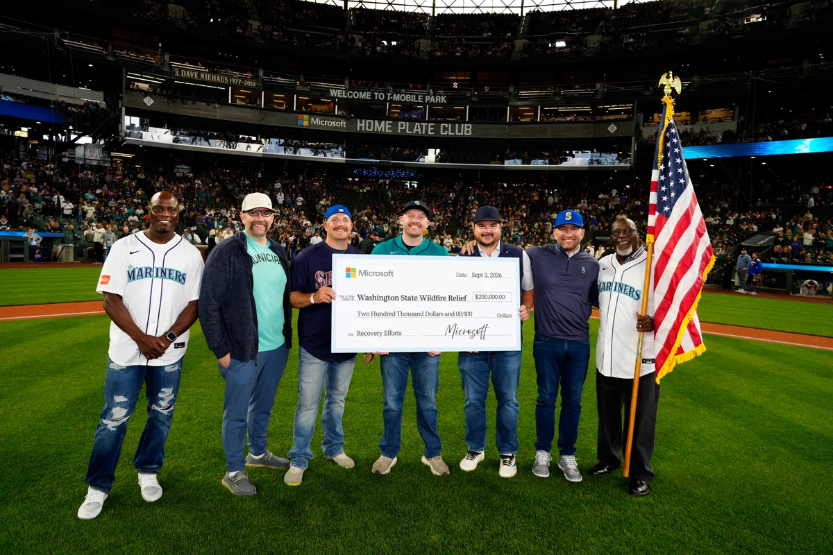 A group of community members and representatives stand on the field at T-Mobile Park before a Seattle Mariners game, holding a ceremonial Microsoft check for $200,000 to support Washington State wildfire relief and recovery efforts. The group is gathered in front of a packed stadium crowd, with two individuals wearing Mariners jerseys and one holding a U.S. flag. The large check, dated September 3, 2026, highlights Microsoft's donation to wildfire recovery efforts in Eastern Washington.