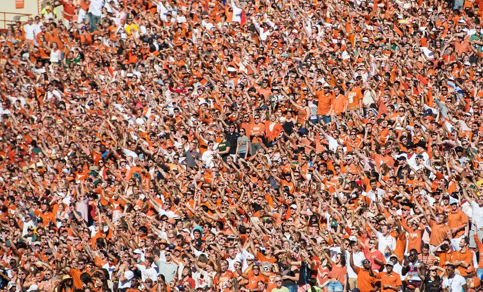 A massive crowd of people, predominantly wearing orange, cheer enthusiastically in a stadium. Many are raising their hands to form the \"Hook 'em Horns\" gesture, creating a sea of orange and hands. Some shirts feature the word \"TEXAS.\"