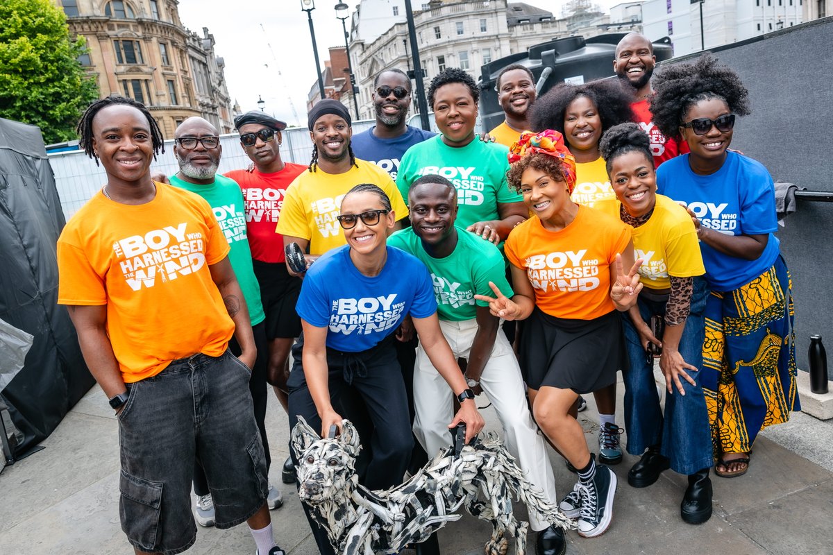 A group of people smiling and wearing colourful "The Boy Who Harnessed the Wind" T-shirts are gathered together outdoors. Some are crouching, others are standing, and they are surrounded by an urban setting with buildings and trees in the background. The atmosphere is lively and cheerful.