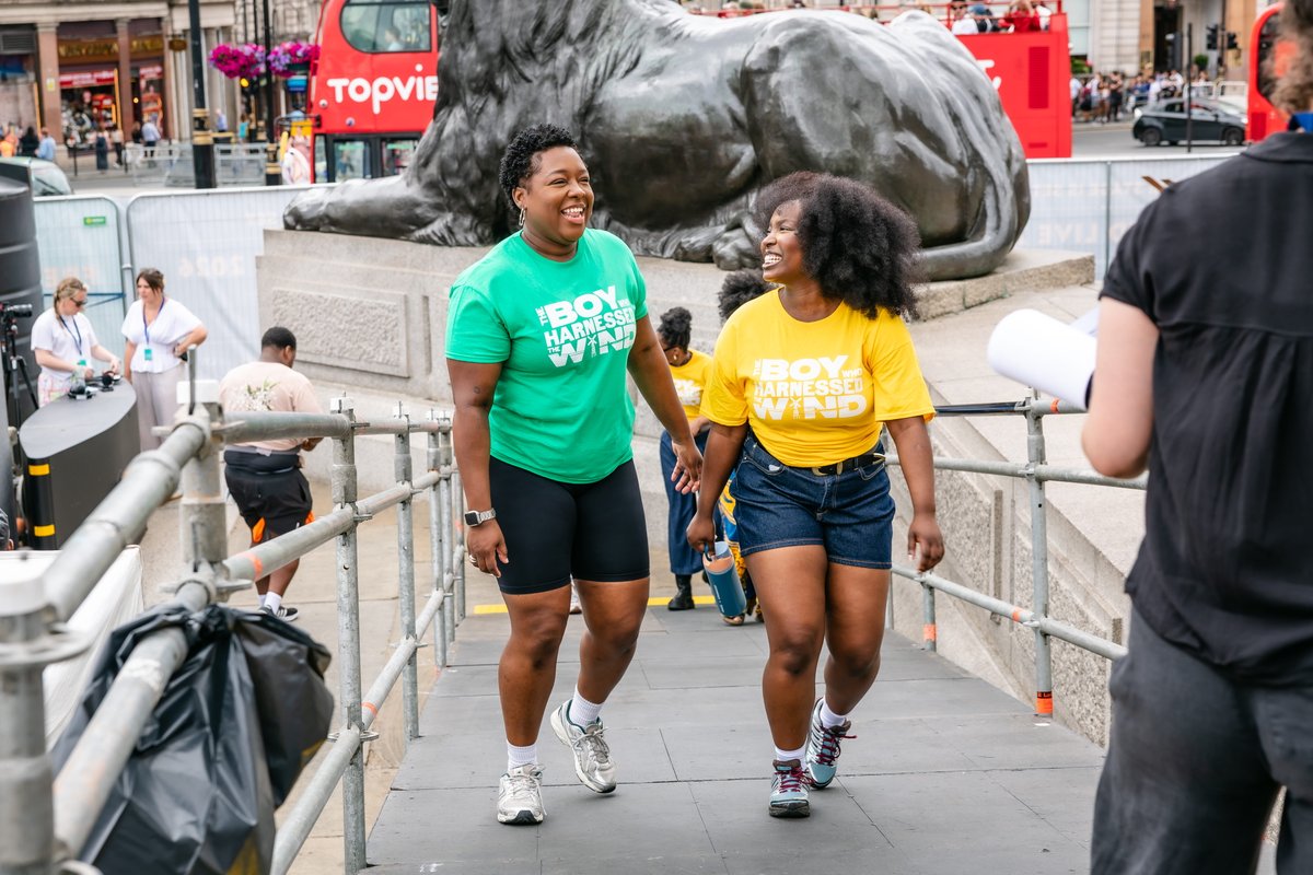 Two people are walking and smiling in front of one of the large lion statues in Trafalgar Square, London. They are wearing brightly colored shirts with text on them. The background includes people and part of a bus.