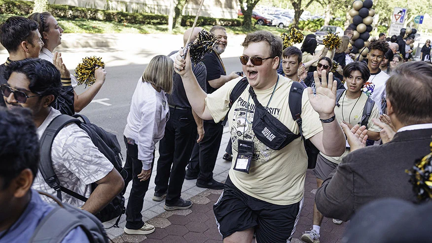 A group of people, many holding gold and black pom-poms, line a pathway in support. One person is in front, enthusiastically engaging with the crowd. Several wear shirts with "Purdue University" visible, and there are black and gold balloons in the background.