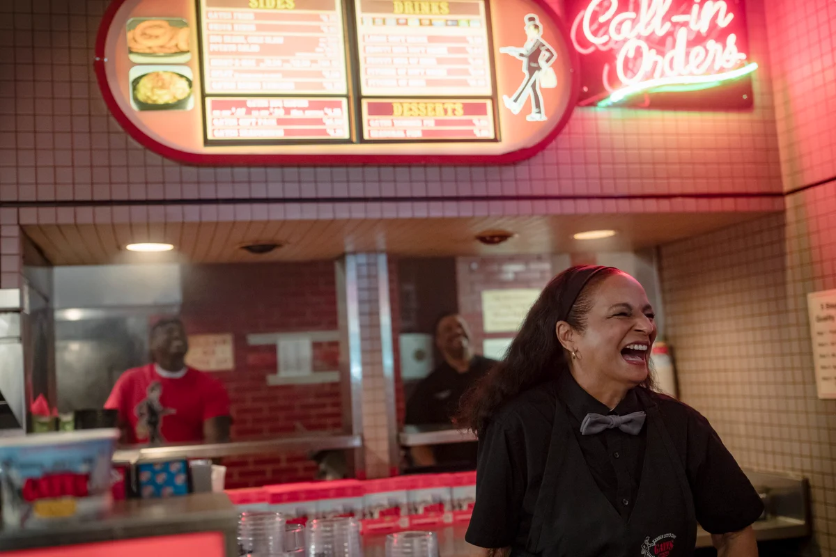 From Ted Lasso. Employees at a fast food restaurant laugh behind the counter and in the kitchen. Neon signs are displayed above where the register is, with menu food items and the text, “Call-in Orders,” shown.