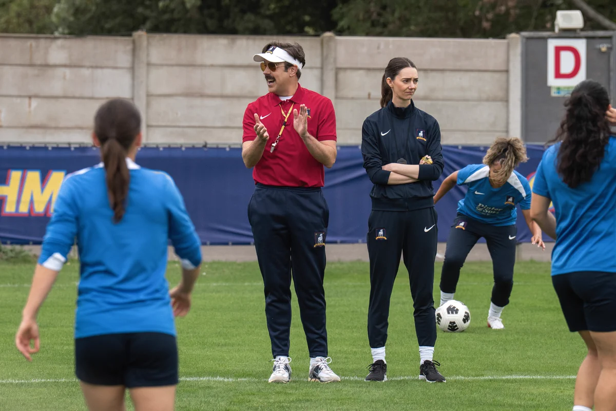 From Ted Lasso. Jason Sudeikis as Ted and Tanya Reynolds as Coach Chilton stand in the middle of practice, with players standing around them. Jason is in mid-clap, as Tanya stands with her arms crossed.