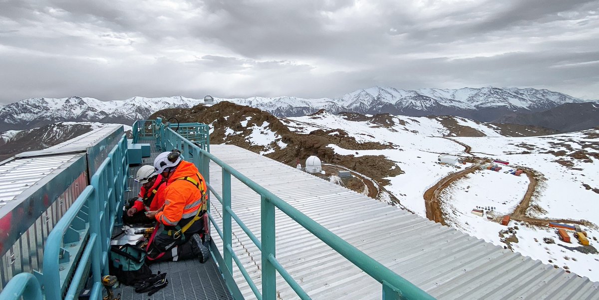Two workers in safety gear doing maintenance work on top of Rubin's silver dome, overlooking snowy mountain peaks under a cloudy sky.