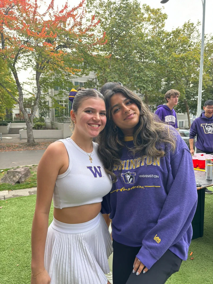 Two young women smile outdoors. One wears a white crop top with a purple 'W' and a pleated skirt. The other wears a purple 'WASHINGTON HUSKIES' sweatshirt. Background shows more people in purple team gear, red cups on a table, and trees.