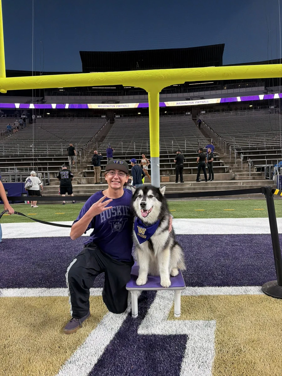 A young man, wearing a \"HUSKIES\" shirt and cap, kneels on a purple and gold football field next to a husky dog sitting on a bench. The man makes a peace sign, smiling. The husky wears a \"W DUBS\" bandana. In the background are stadium stands, a yellow goalpost, and a \"WASHINGTON FOOTBALL\" sign.
