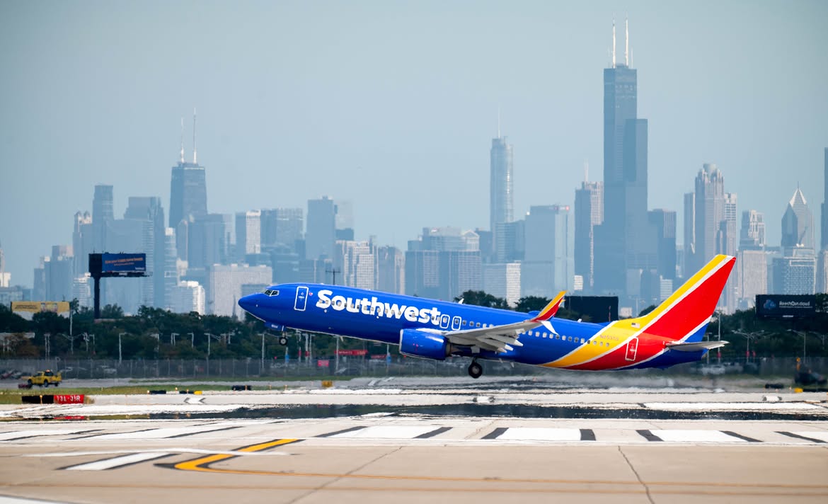 A Southwest Airlines plane takes off from a runway with the Chicago skyline visible in the background. The sky is clear, and the cityscape includes several tall buildings.