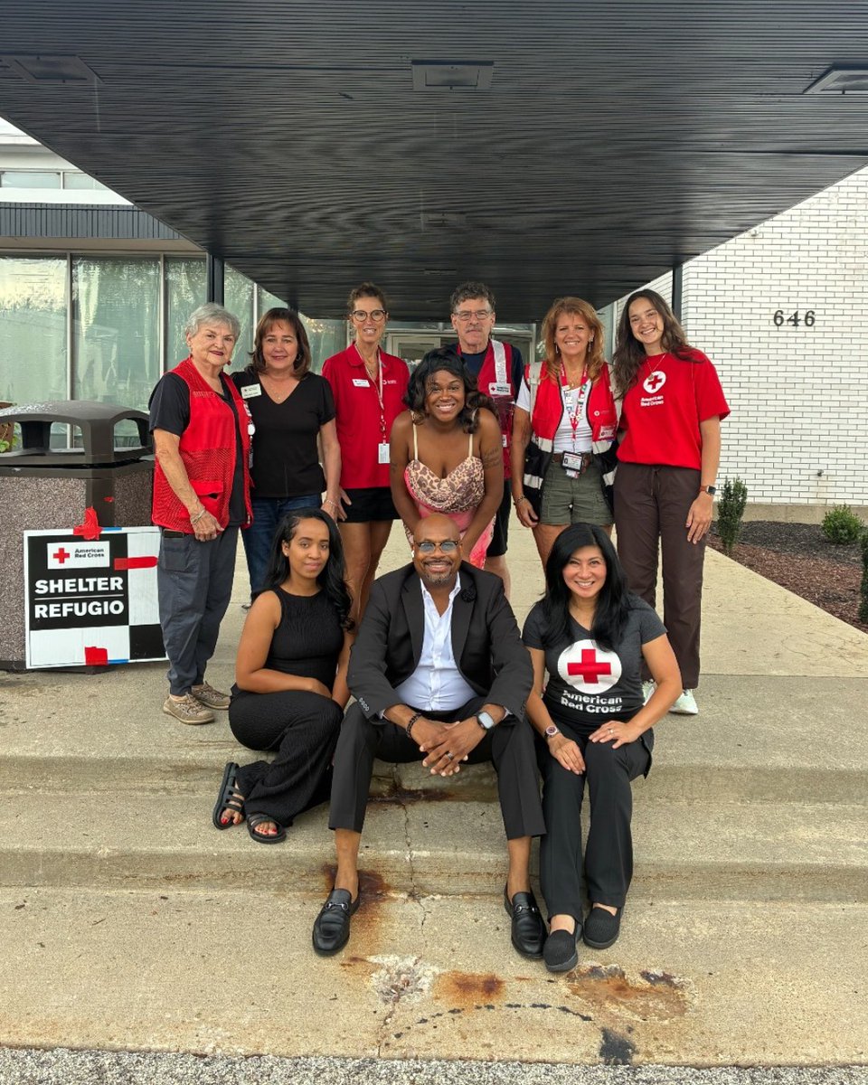 A group of individuals is gathered in front of a building entrance with a sign that reads "Shelter, Refugio." They are dressed in casual attire, some wearing Red Cross vests or shirts. The group appears to be smiling, and the setting suggests an organized event or volunteer gathering.