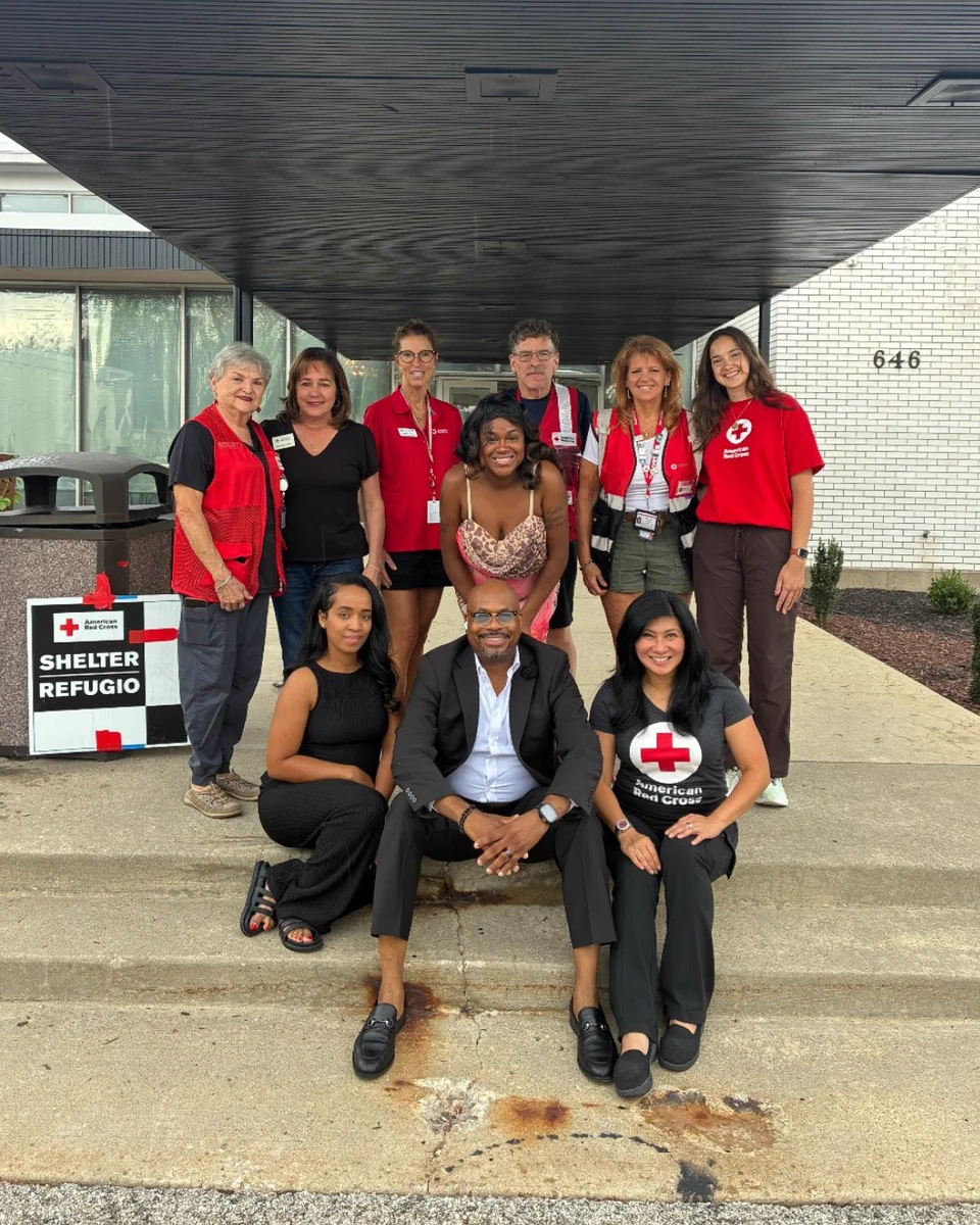 A group of individuals is gathered in front of a building entrance with a sign that reads "Shelter, Refugio." They are dressed in casual attire, some wearing Red Cross vests or shirts. The group appears to be smiling, and the setting suggests an organized event or volunteer gathering.