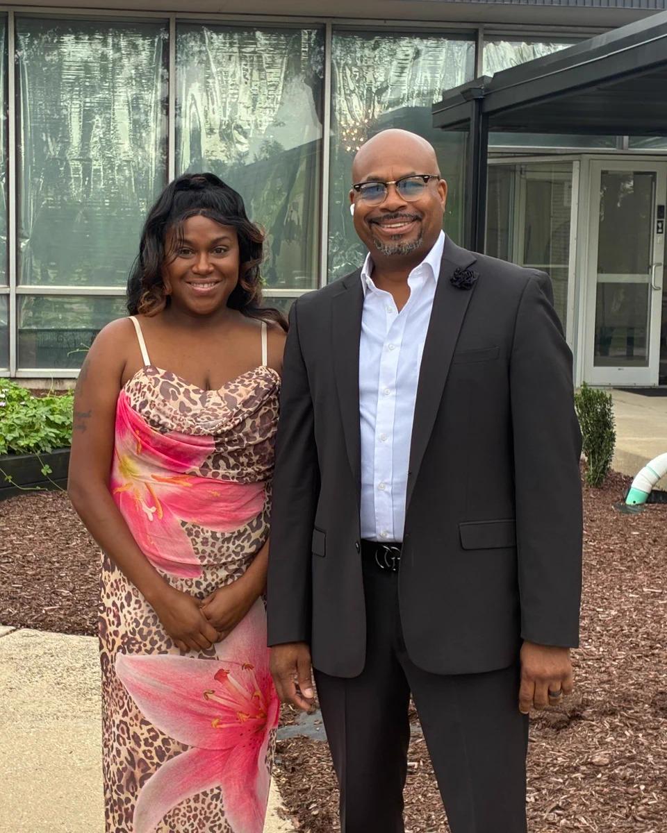 Two people are standing outside a modern building with large glass windows. One is in a floral and leopard print dress, and the other is in a dark suit with a white shirt. Both are smiling. The backdrop includes some greenery and landscaping.