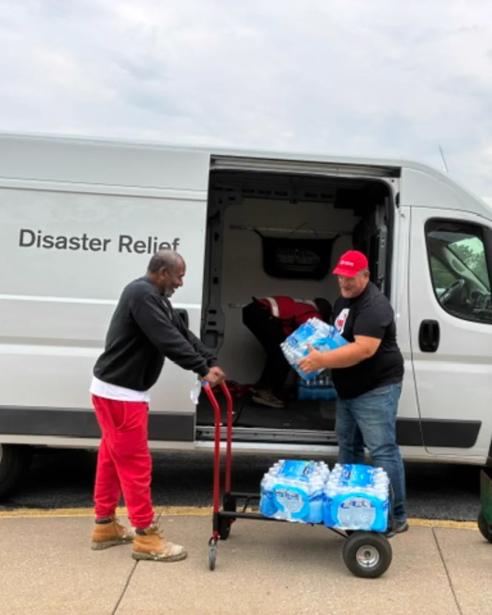 A person is handing bottled water to another individual outside an American Red Cross disaster relief van. They are using carts to assist with the transportation of the water. The scene takes place on a paved area with a cloudy sky in the background.