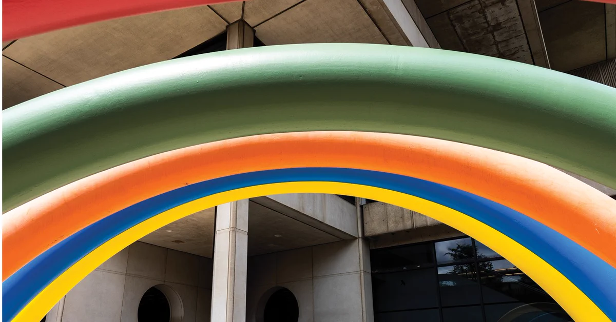 A close-up view of a colorful art installation featuring large, curved arches in red, green, orange, yellow, and blue. The concrete exterior of a building is visible in the background, and the scene takes place outdoors.