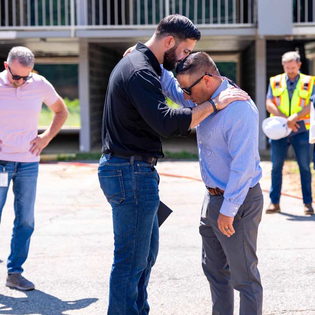 Two men share a comforting embrace outdoors, one resting his forehead on the other's head in a moment of support.