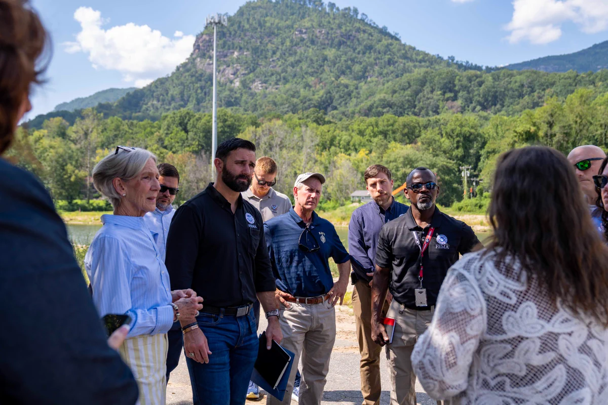 Group of diverse adults engaged in an outdoor discussion with a lush green mountain and blue sky in the background.