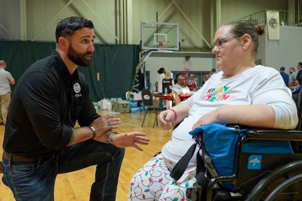 FEMA Administrator kneeling and talking attentively to a woman in a wheelchair inside a gymnasium.
