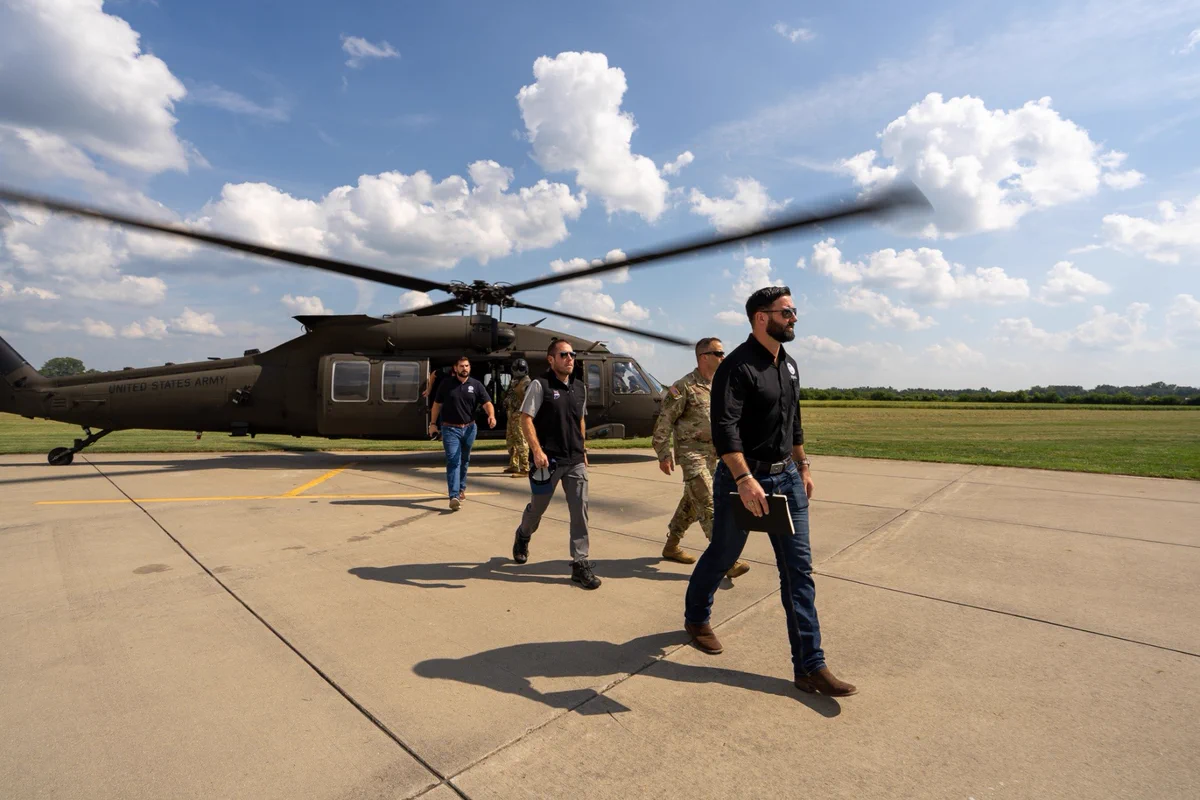 Four men, including one in military uniform, walk away from a black helicopter on a sunny airfield with scattered clouds.