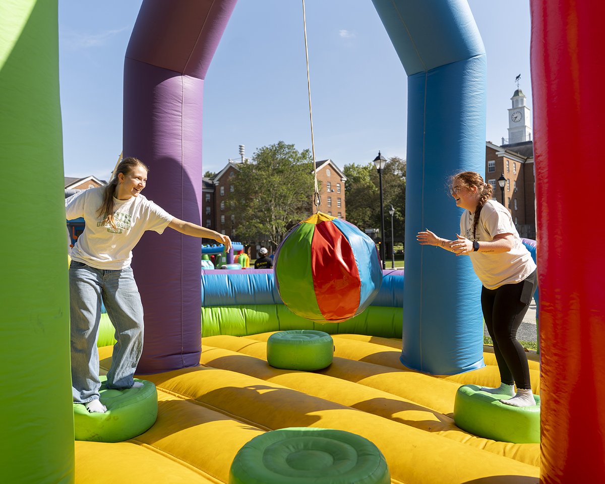 Two individuals play on an inflatable obstacle course outdoors, balancing on green circular platforms while trying to touch a suspended colorful ball. Brick buildings and trees are visible in the background under a clear blue sky.