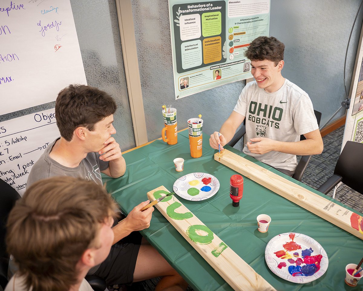 Three individuals are gathered around a table engaged in a paint activity. Two Ohio University Bobcats shirts and a poster about transformational leadership are visible in the background. The table is covered with a green cloth and paint supplies.