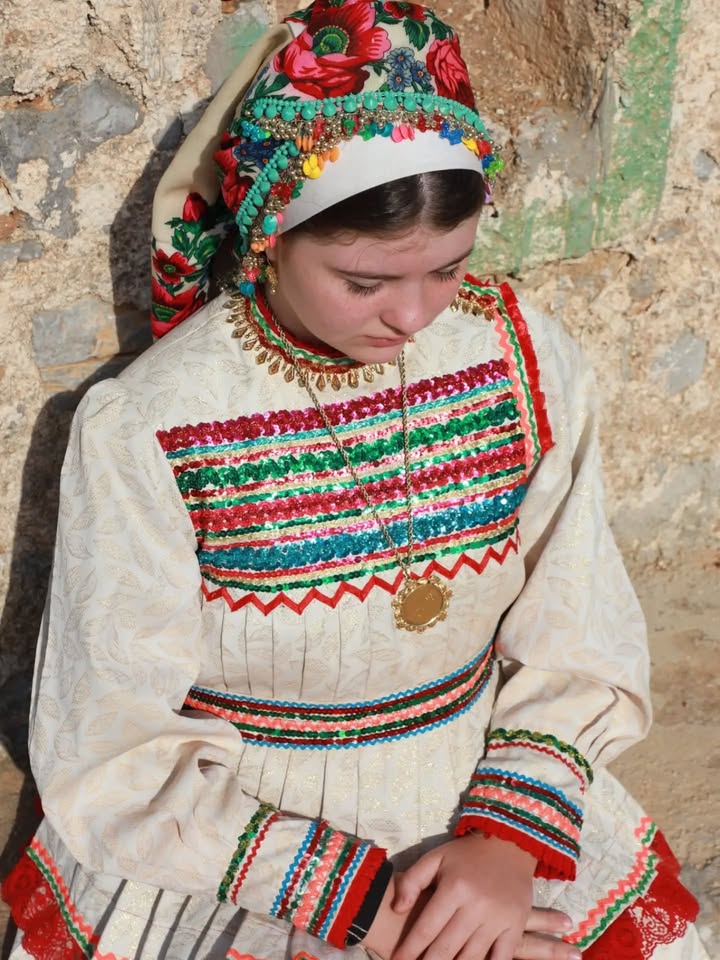A person is wearing traditional clothing with intricate embroidery, featuring colorful patterns and a headdress adorned with flowers and beads. The background shows a textured stone wall.