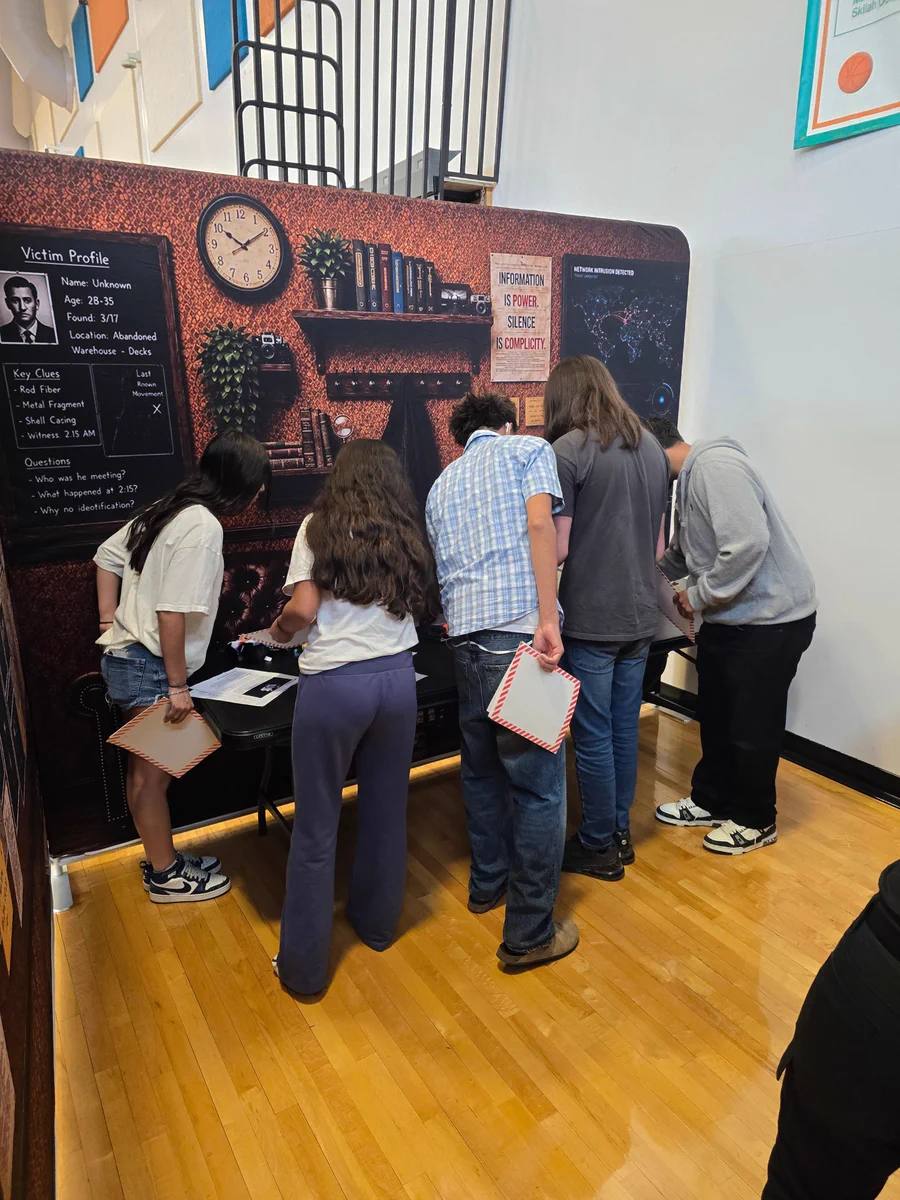 Group of five students closely examining an interactive mystery-solving display on a gymnasium wall.