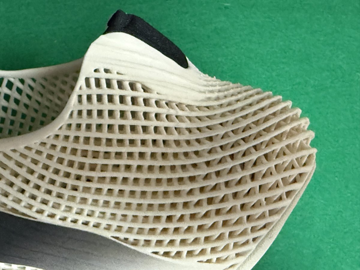 hackin_kun's tweet photo. adidas FUTURECOOL。気になる接合部を観察した。
アッパー〜ミッドのクリーム格子はAMの一体構造。一方、黒の伸縮部・サイドパッチ・トウ・アウトソールゴムは、格子セルへ流し込んだ共成形ではなく、ポストキュア後の接着にみえる。
アウトソールは黒ゴム単体ではなく、クリームのキャリアと黒を先に一体化した成形ユニットを骨格へ貼ってる感じ。先端の大きな巻き上げも、平面シートの折り返しではなく、プリント曲面に合わせた立体シェルに仕上げて貼り合わせている感じ。
公表の”fusing resin with specialised materials”は化学的な共キュアというより、幾何の嵌合＋接着による機能融合、と考えたほうがしっくりくる。
黒い部分が伸びるから レース無しのClimacoolより履きやすい。来春発売の時には、Climacoolの時のように軽量化などがされるとよいと思う。