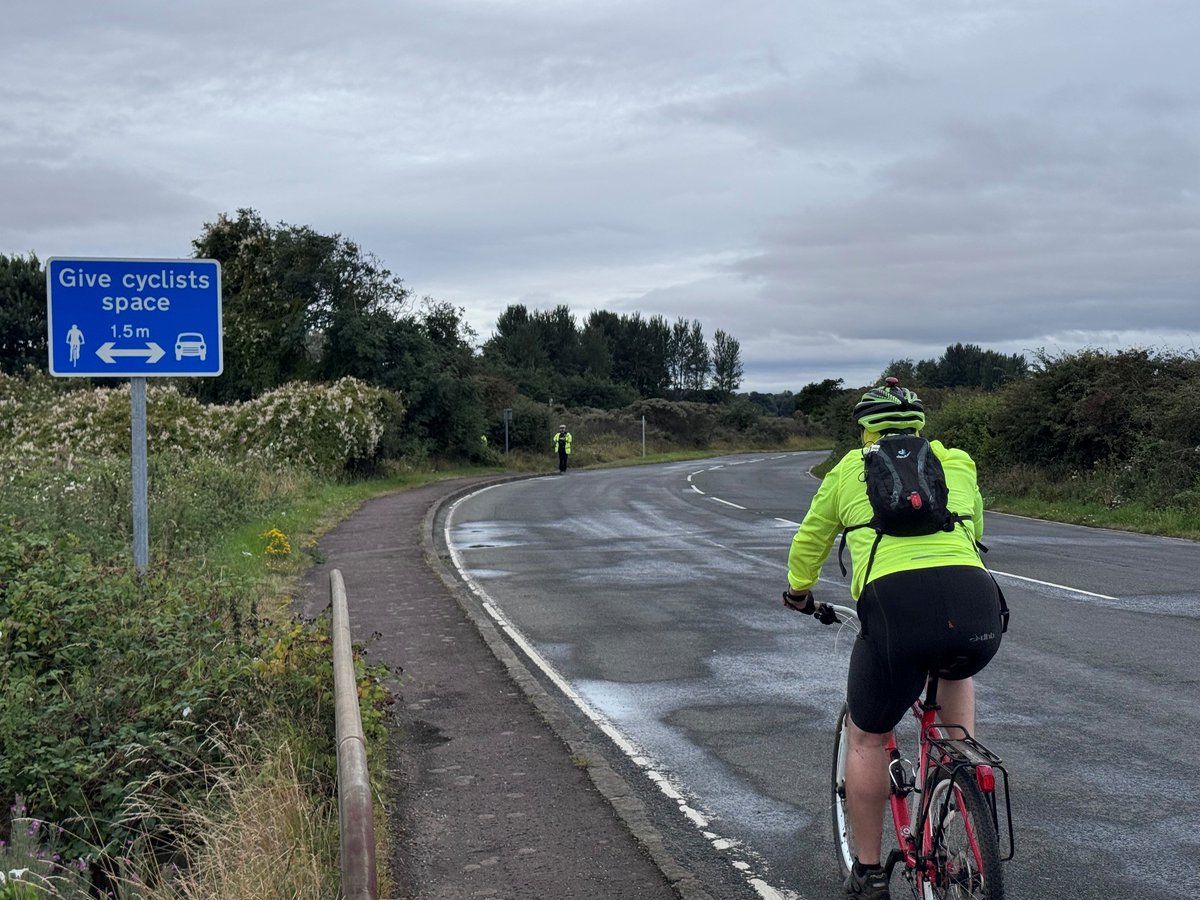 A cyclist in a neon yellow jacket rides along a rural road past a 'Give cyclists space' sign.
