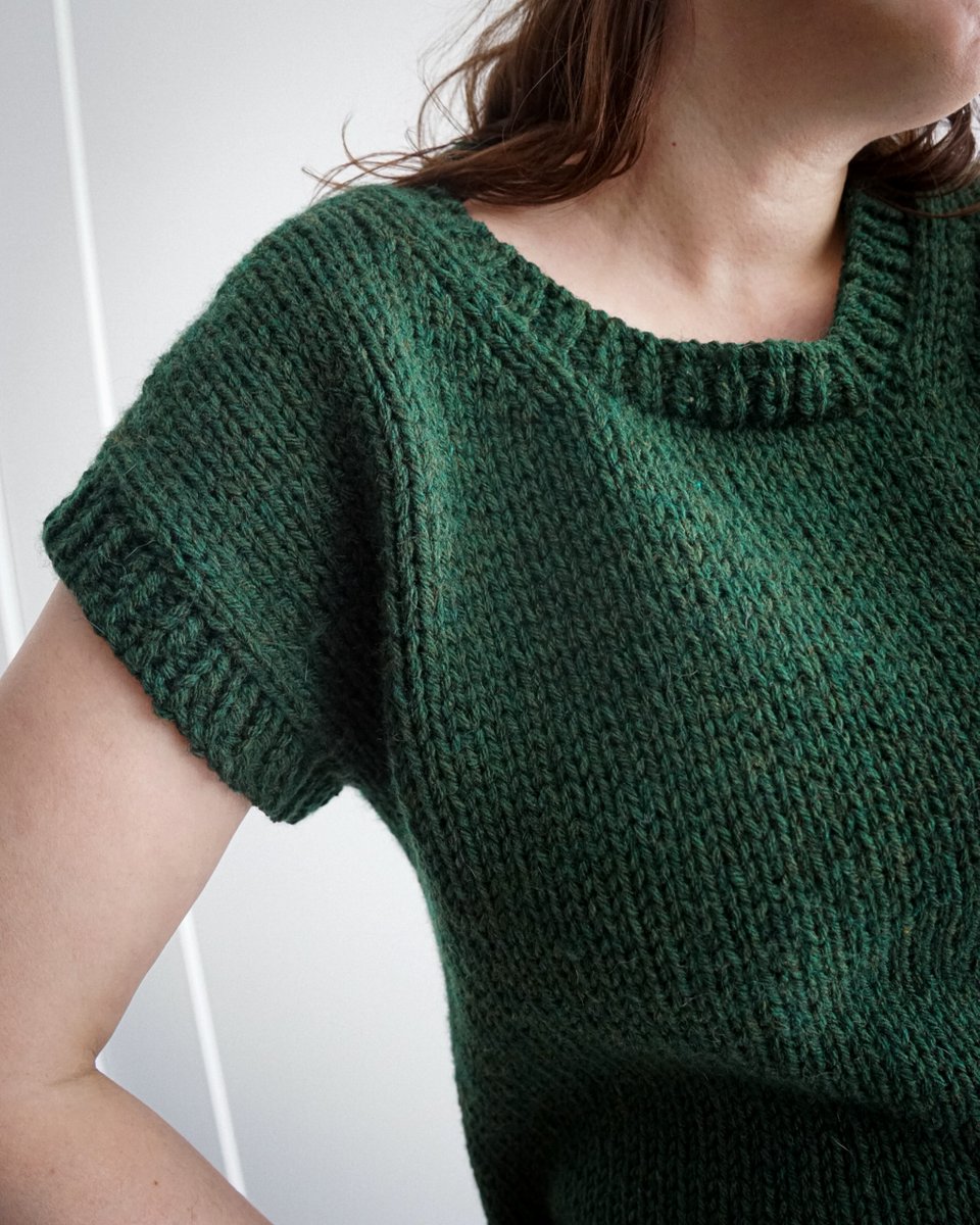 Close-up of a person wearing a hand-knitted, dark green short-sleeve sweater with a ribbed neckline and cuffs.