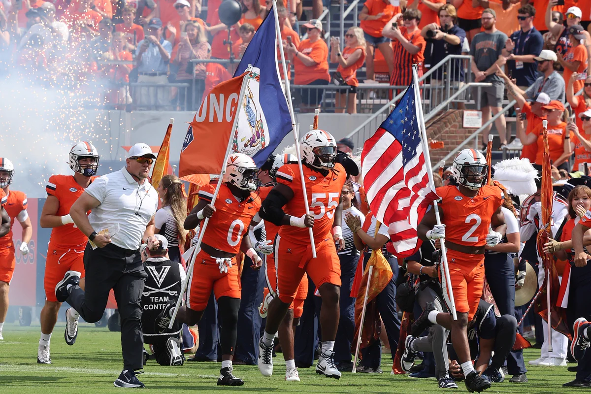 A group of UVA football players running onto the field at Scott Stadium, led by one player holding a United States flag and another carrying a UVA flag. In the background, a crowd of fans in orange attire watch, while smoke and sparks create an energetic atmosphere.
