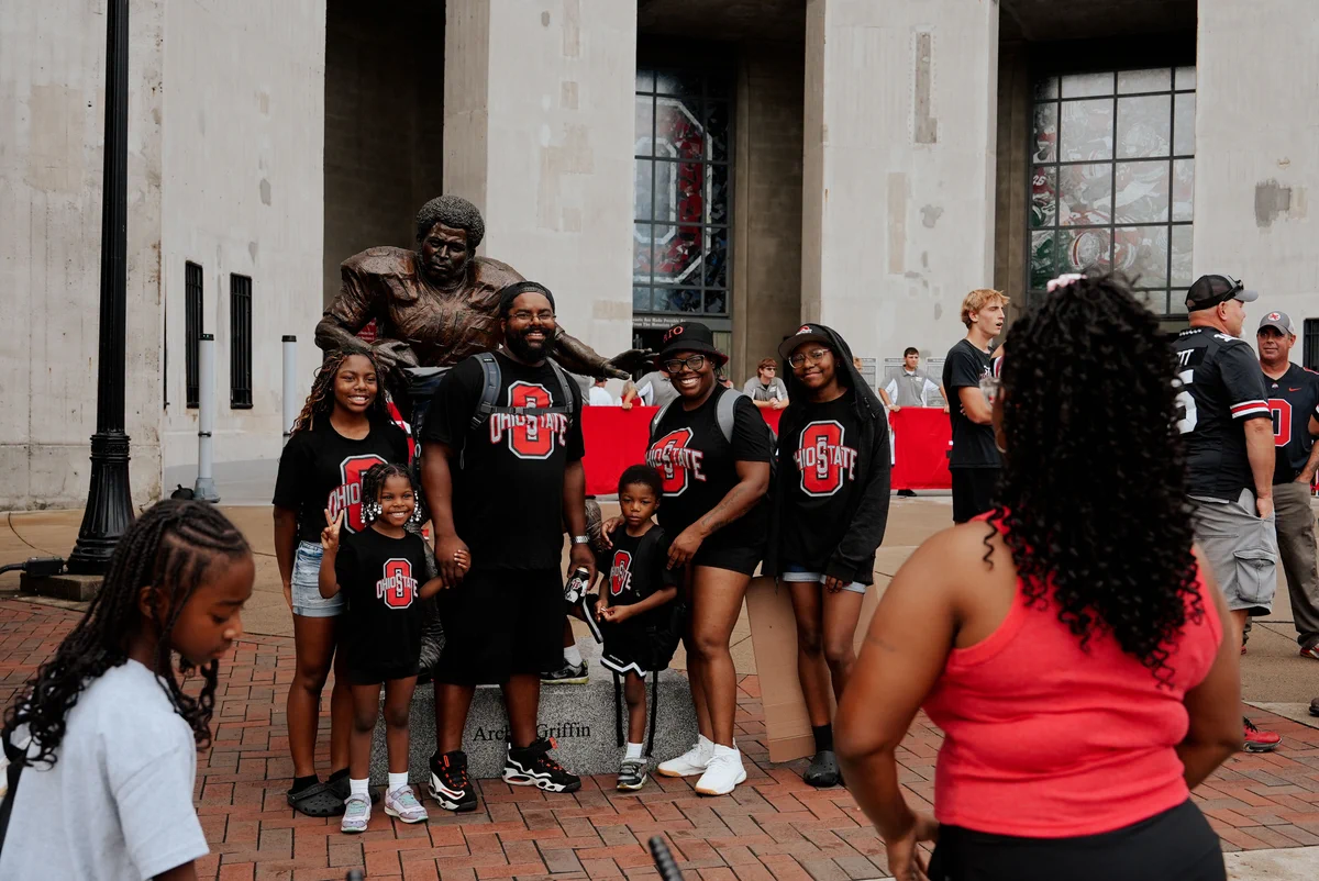 A group of people wearing Ohio State shirts pose in front of a statue at Ohio Stadium. A person stands in the foreground taking their photo. Other visitors and the stadium exterior are visible in the background.