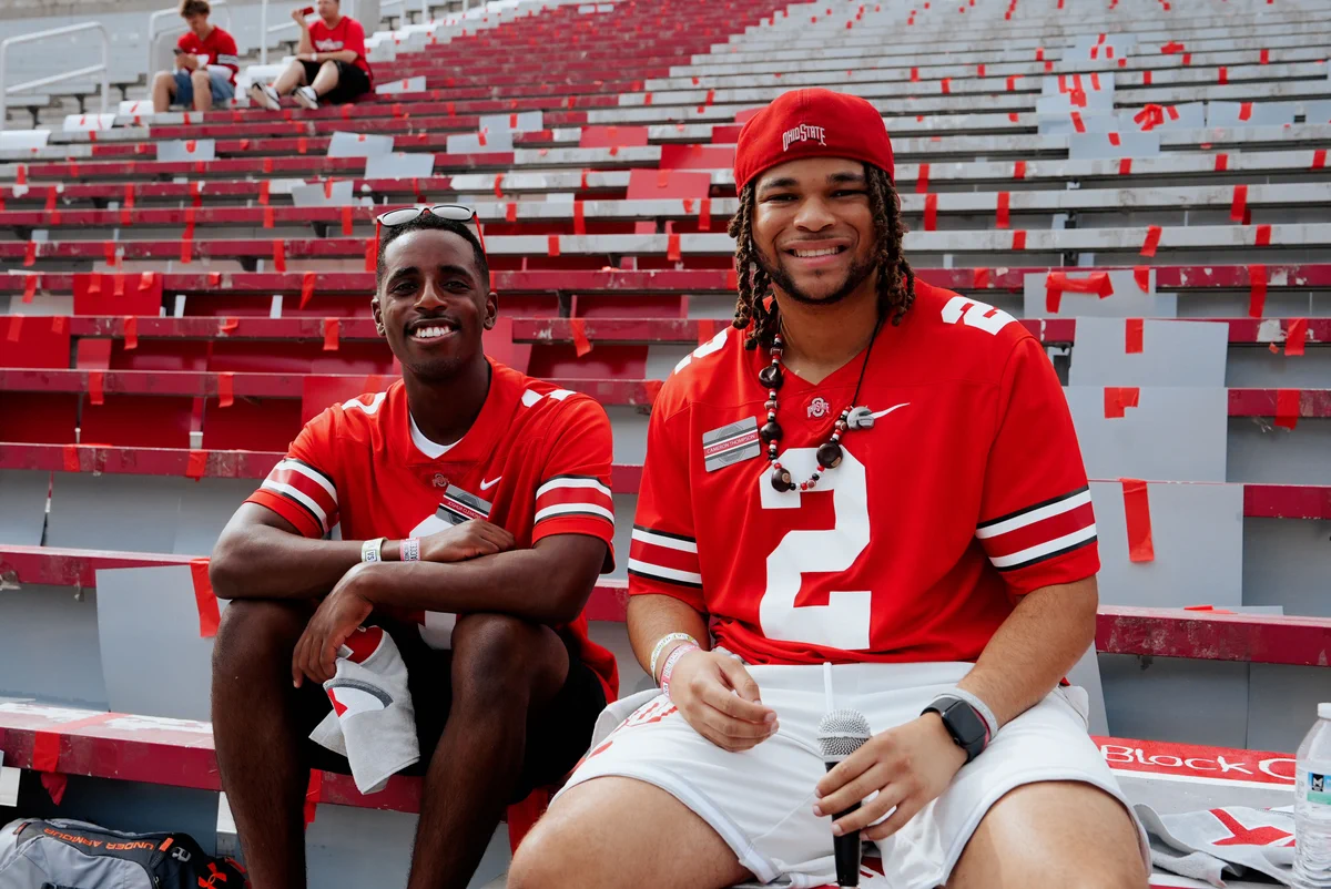 Two individuals sit in stadium bleachers, wearing red Ohio State jerseys. The seats have red and white stripes. They appear relaxed and are smiling.