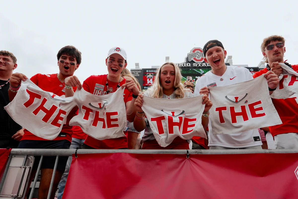 A group of enthusiastic Ohio State fans stand at a stadium barrier, holding towels with the word "THE" printed in red. They are cheering energetically. The Ohio State University logo is visible in the background.