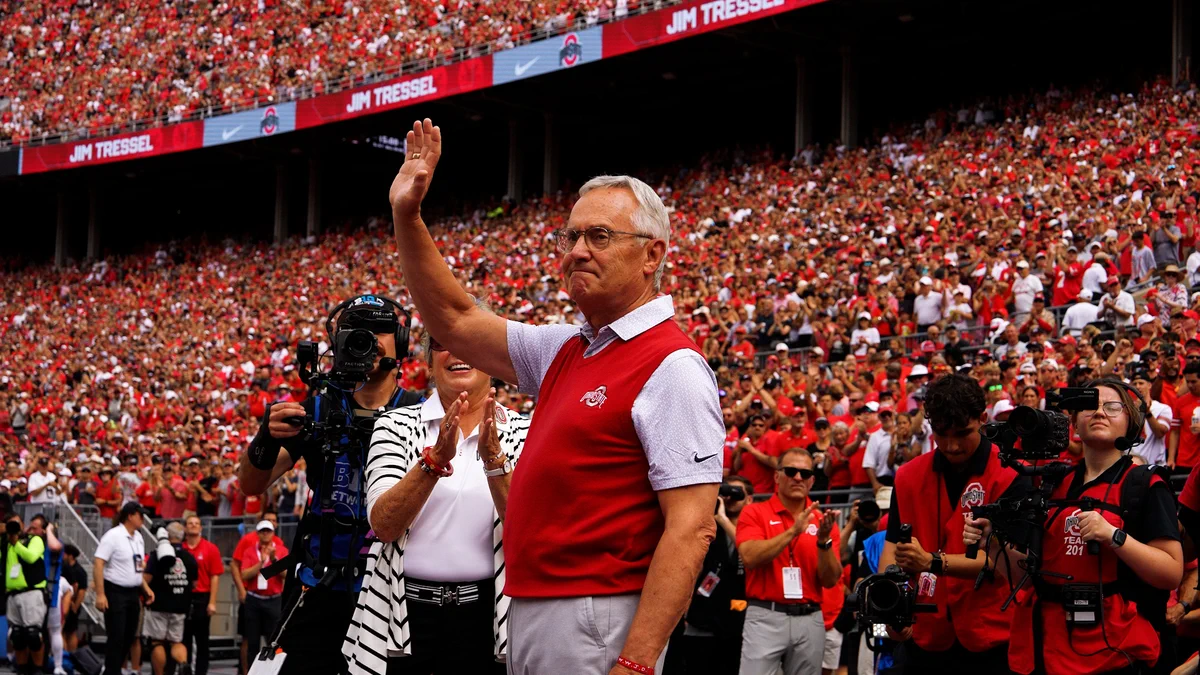 A person is waving to a large crowd at a sports stadium. The audience is filled with people wearing red and white. A banner above displays the name "Jim Tressel." Camera operators and photographers are visible in the foreground.