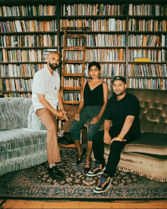 A group of three people sitting in front of a large bookshelf filled with books. The setting includes a beige sofa, a gray striped couch, and a patterned rug.