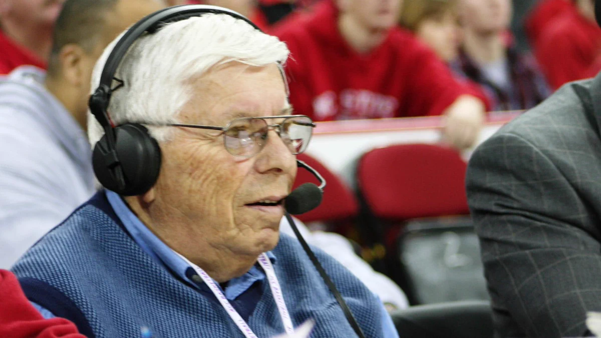 Elderly man with white hair wearing glasses and a headset, speaking during a live sports broadcast at an indoor event.