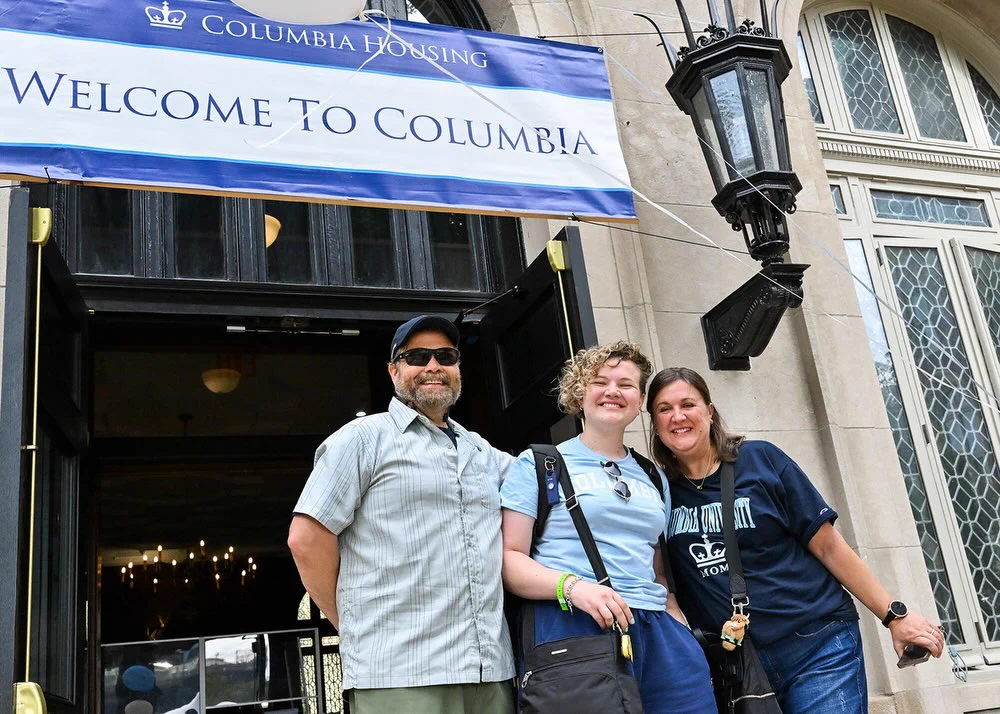 A group of three people smiling in front of a building with a "Welcome to Columbia" banner. The entrance has large windows and ornate lanterns. The group appears content and happy.