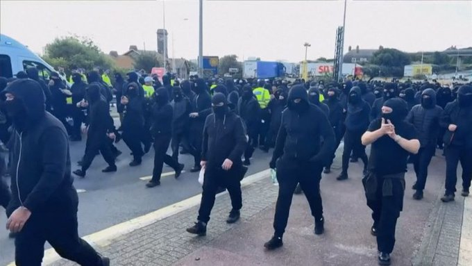 YourAnonCentral's tweet photo. A Russian &amp; Israeli-funded disruption operation sees black-clad right-wing thugs in balaclavas block UK’s largest international ferry port, which handles roughly one-third of UK–EU trade. An estimated £16.4 million worth of trade per hour. Promoted by IDF linked Tommy Robinson. https://t.co/uZUiVNHbTs