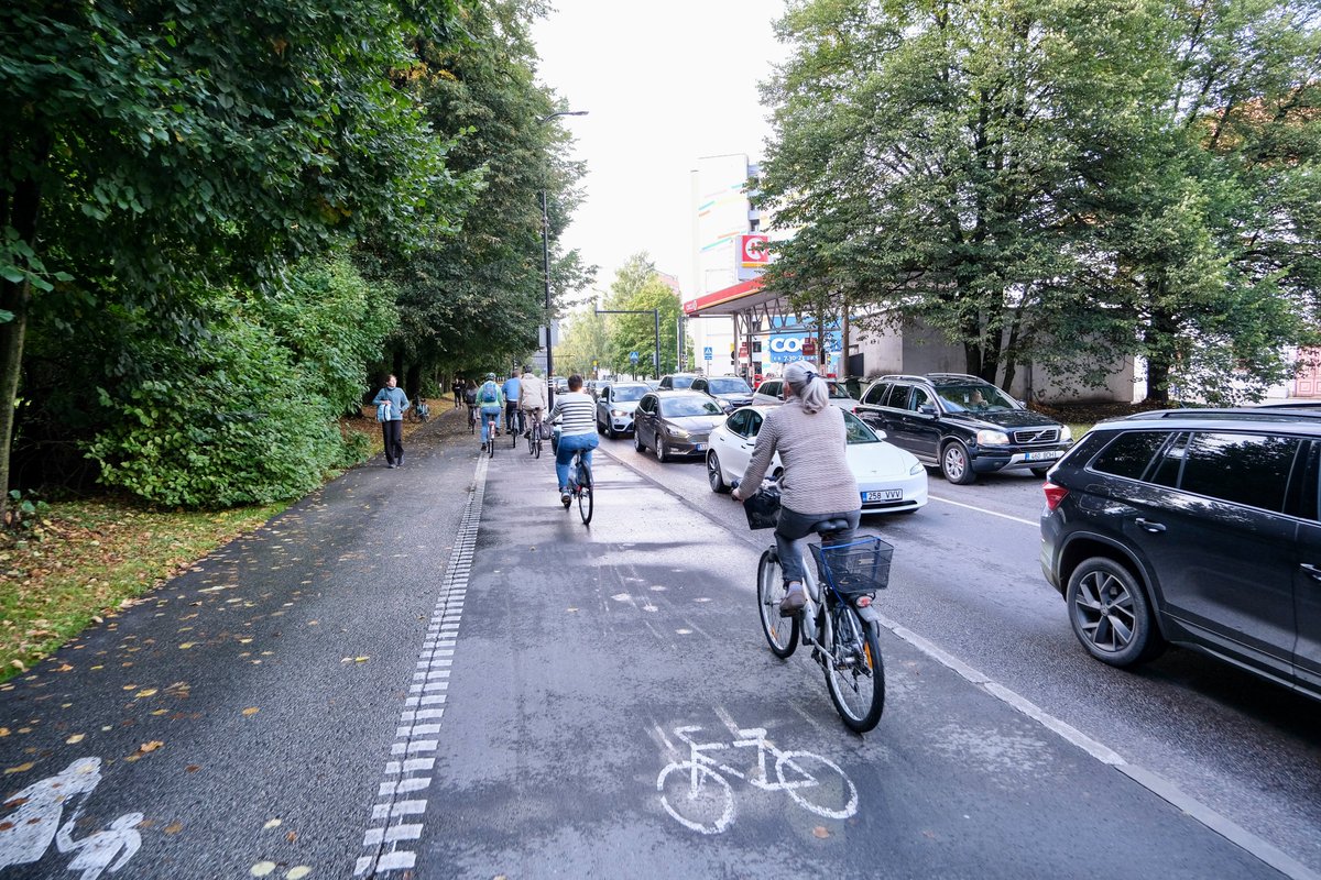 Cyclists in casual autumn attire ride along dedicated cycling infrastructure lined with buildings and trees on the streets of Tallinn, Estonia.