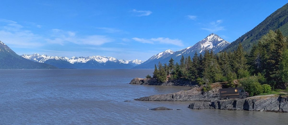 Snow-capped mountains surround a calm body of water, with a forested rocky shoreline and a UPS truck parked on the right.