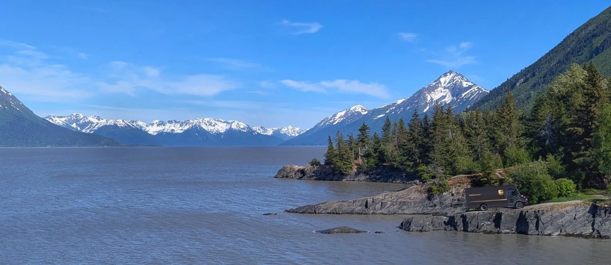 Snow-capped mountains surround a calm body of water, with a forested rocky shoreline and a UPS truck parked on the right.